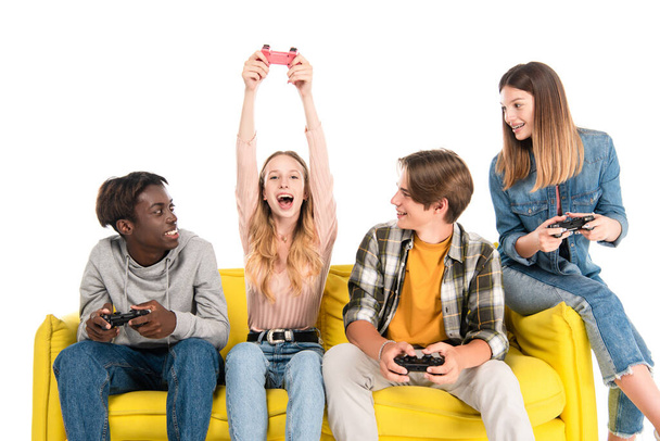 KYIV, UKRAINE - AUGUST 21, 2020: Cheerful multicultural teenagers playing video game on yellow sofa isolated on white - Fotoğraf, Görsel