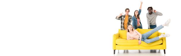 Панорамный снимок жизнерадостных мультиэтнических подростков, показывающих знак мира возле желтого дивана на белом фоне - Фото, изображение