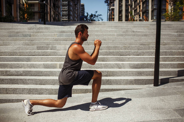 Young man exercising outside. Side view of guy athletist sitting in one leg squat position holding hands together in front himself. Training or having workout. - Photo, Image