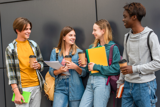 Позитивные подростки с блокнотами и кофе будут стоять возле здания на открытом воздухе - Фото, изображение