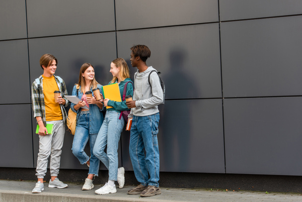 Mosolygó többnemzetiségű tinédzserek hátizsákokkal, jegyzetfüzetekkel és kávéval az épület közelében  - Fotó, kép