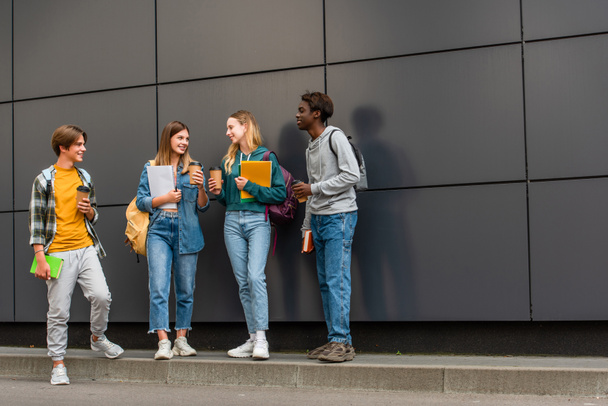 Adolescentes multiétnicos positivos con cuadernos y café para ir a hablar cerca del edificio al aire libre  - Foto, imagen