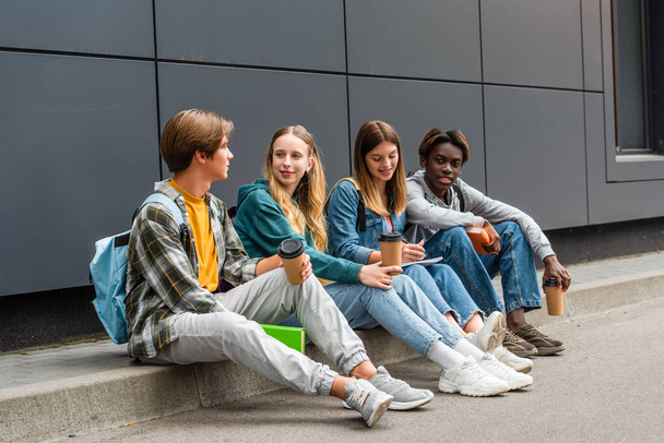 Adolescentes multiétnicos positivos con café para llevar y libros sentados en la acera cerca del edificio  - Foto, Imagen
