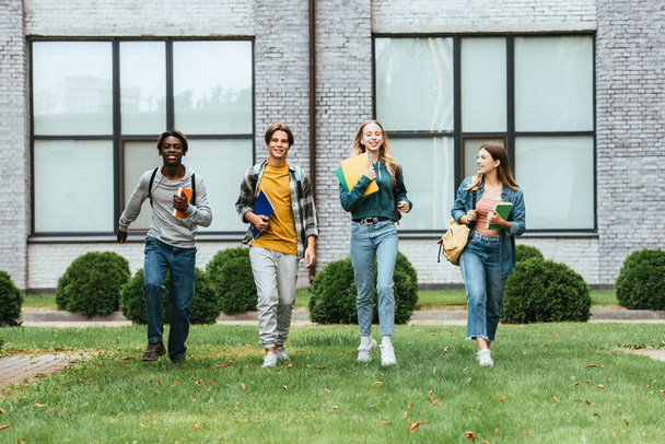 Вибірковий фокус веселих багатоетнічних підлітків з блокнотами та рюкзаками, що ходять по траві на відкритому повітрі
  - Фото, зображення