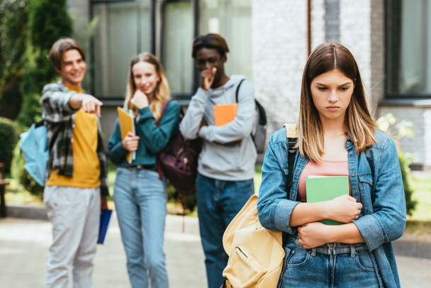 Selektywne skupienie smutnej nastolatki z książką i plecakiem stojącej w pobliżu wieloetnicznych uczniów wskazujących palcem na zewnątrz  - Zdjęcie, obraz