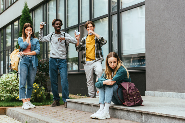 Selektiver Fokus eines aufgebrachten Mädchens in der Nähe lächelnder multikultureller Teenager mit klumpigem Papier im Freien  - Foto, Bild