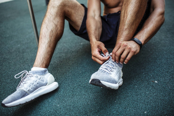 Young man exercising outside. Cut view of sportsman sitting on fround and tie shoe laces. Preparing for exercising. Slim well built muscled legs and calfs. - Photo, image