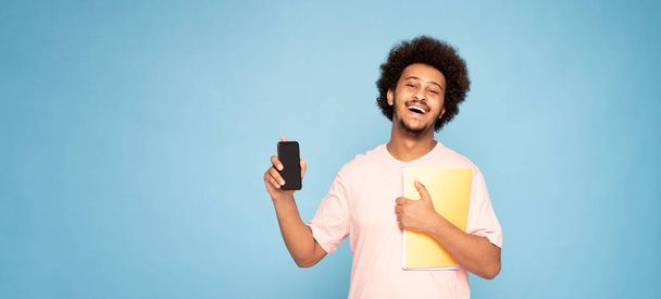 Cheerful young african guy in casual t-shirt holding mobile phone and notebook, smiling. - Fotoğraf, Görsel
