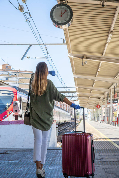 Widok z tyłu nierozpoznawalnej kobiety w lateksowych rękawiczkach ciągnącej walizkę i sprawdzającej czas spaceru po peronie stacji kolejowej podczas epidemii - Zdjęcie, obraz