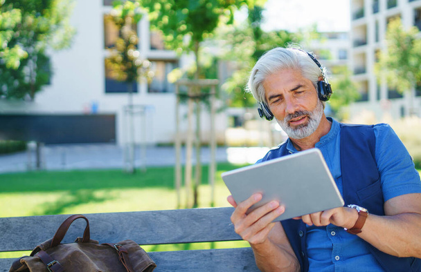 Portrait of mature man with headphones sitting outdoors in city, using tablet. - Photo, Image