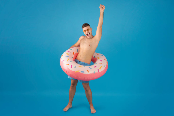 Full length portrait of handsome millennial man wearing inflatable ring and making YES gesture over blue background - Photo, Image