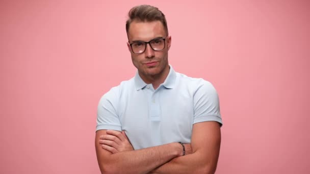 sexy joven hombre casual con los brazos cruzados está usando gafas, mirando a la cámara y sonriendo en el fondo rosa - Imágenes, Vídeo