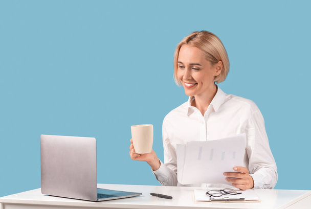 Femme d'affaires positive travaillant avec des documents et buvant du café devant un ordinateur portable au bureau, fond bleu - Photo, image