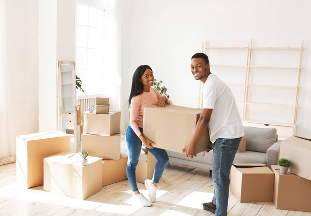 Positive black guy with his girlfriend holding cardboard box in their new house on moving day - Photo, Image
