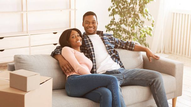 Glückliches, müdes schwarzes Paar, das es sich nach einem langen Umzugstag in seiner neuen Wohnung auf dem Sofa gemütlich macht. Panorama - Foto, Bild