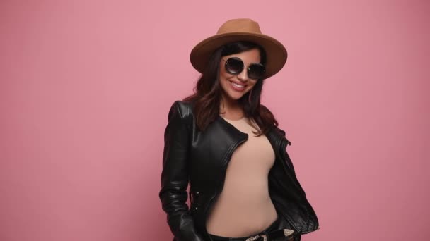 sexy menina em jaqueta de couro vestindo chapéu e óculos de sol sorrindo, de mãos dadas em bolsos, tocando chapéu, olhando para baixo e posando no fundo rosa - Filmagem, Vídeo