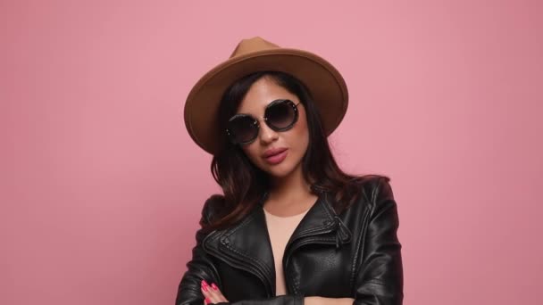 sexy donna fresca in giacca di pelle indossa cappello e occhiali da sole, braccia incrociate e posa, sorridente, tenendo le mani in tasca e in piedi su sfondo rosa - Filmati, video