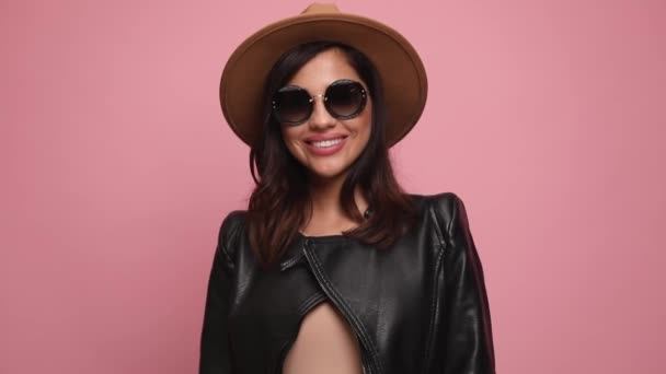 szczęśliwa kobieta w skórzanej kurtce nosząca okulary przeciwsłoneczne i kapelusz, uśmiechnięta, układająca włosy, patrząca w dół i pozująca na różowym tle - Materiał filmowy, wideo