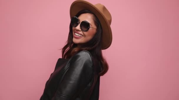menina legal em jaqueta de couro vestindo óculos de sol e chapéu, sorrindo, posando, fazendo um gesto de coração, se divertindo e dançando em fundo rosa - Filmagem, Vídeo