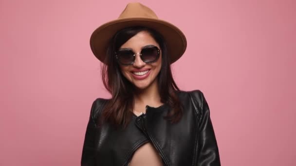 happy young girl in leather jacket smiling, sticking out tongue and throwing hat in the air, walking on pink background - Footage, Video