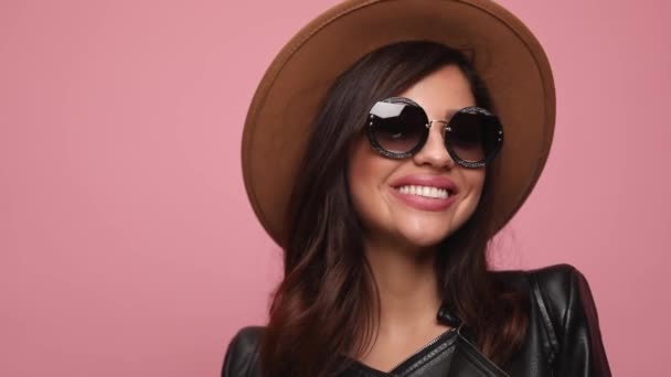 young girl in leather jacket wearing hat and sunglasses smiling, looking to side and posing on pink background - Footage, Video