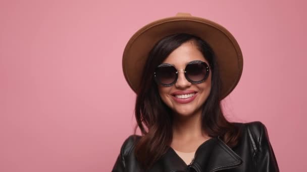 happy girl in leather jacket wearing hat and sunglasses running fingers through hair and fixing it, looking to side and smiling, posing on pink background - Footage, Video