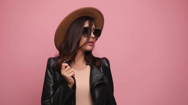 sensual menina legal em jaqueta de couro, vestindo chapéu e óculos de sol, segurando jaqueta, olhando para o lado e posando, de mãos dadas em bolsos no fundo rosa - Filmagem, Vídeo