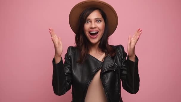 surprised cool girl in leather jacket opening mouth, holding hands in the air, covering mouth with hands, dancing and celebrating victory, smiling on pink background - Footage, Video