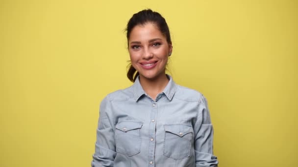 casual young woman in blue denim shirt crossing arms, smiling, posing and standing on yellow background - Footage, Video