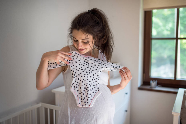 Portrait of pregnant woman indoors at home, holding girls bodysuit. - Foto, afbeelding