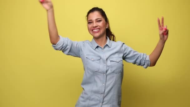 mujer joven en camisa de mezclilla azul haciendo gestos de baile, señalando con los dedos, sosteniendo los puños en el aire, bailando y celebrando sobre fondo amarillo - Metraje, vídeo