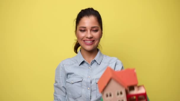 sonriente chica casual joven en camisa de mezclilla azul mostrando y presentando modelo de edificio, haciendo pulgares hacia arriba signo sobre fondo amarillo - Imágenes, Vídeo