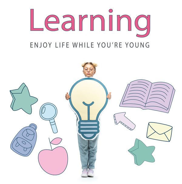 full length view of schoolchild in eyeglasses holding paper cut light bulb near learning, enjoy life while you are young lettering on white - 写真・画像