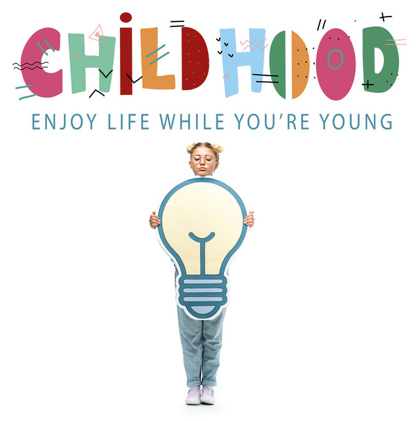 full length view of schoolchild in eyeglasses holding paper cut light bulb near childhood, enjoy life while you are young lettering on white - Foto, Imagen