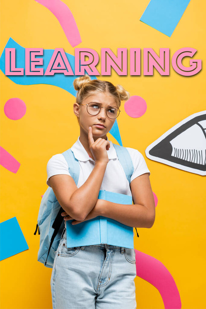 thoughtful schoolgirl in eyeglasses looking away while holding book near paper pencil, colorful elements and learning lettering on yellow - Foto, Imagem