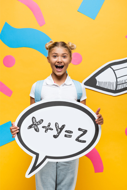 excited schoolchild holding speech bubble with math formula illustration near paper elements and pencil on yellow  - Фото, изображение