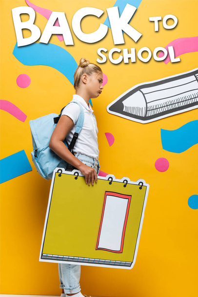 side view of upset schoolgirl holding notebook maquette near back to school lettering, paper pencil and decorative elements on yellow - Photo, Image