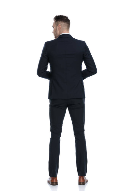 白いスタジオの背景に立っている間に肩を見上げ、ジャケットを調整する神秘的なビジネスマンのバックビュー - 写真・画像