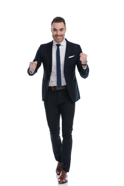 Positiver Geschäftsmann lächelt und feiert mit geballten Fäusten, während er auf weißen Studiohintergrund tritt - Foto, Bild