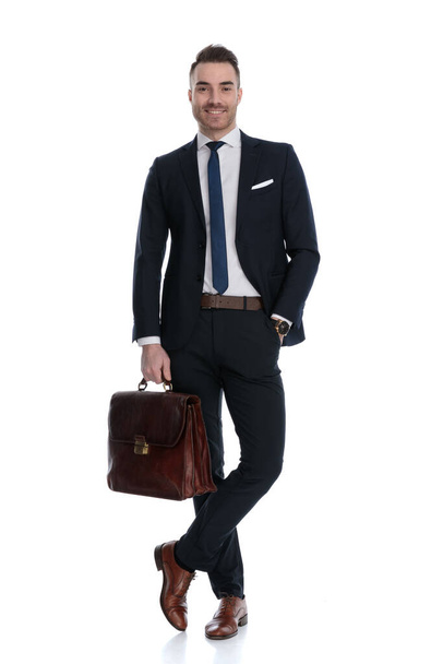 Feliz hombre de negocios sosteniendo el maletín y sonriendo con la mano en el bolsillo mientras está de pie con las piernas cruzadas sobre fondo blanco del estudio - Foto, Imagen