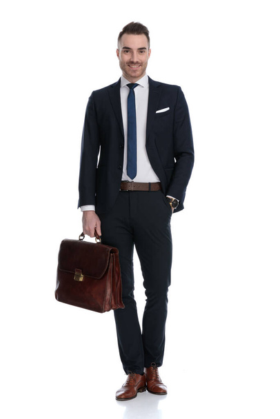 Positive businessman holding briefcase and smiling with hand in pocket while standing on white studio background - Foto, Imagen