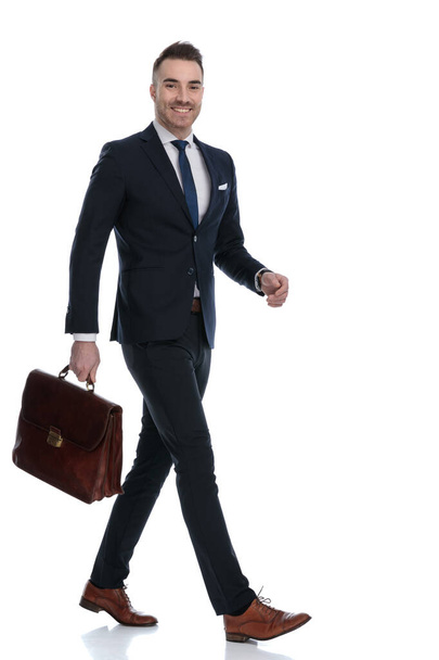 Side view of positive businessman holding briefcase and smiling while walking on white studio background - 写真・画像