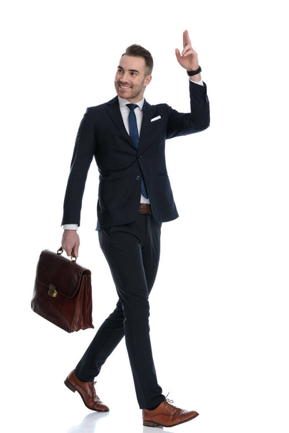 Vista lateral del feliz hombre de negocios sosteniendo el maletín y saludando mientras camina sobre fondo blanco del estudio - Foto, imagen