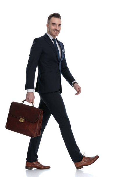 Side view of businessman holding briefcase and smiling while moving on white studio background - Zdjęcie, obraz