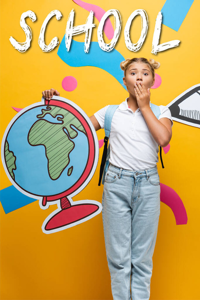 shocked schoolgirl covering mouth while holding globe maquette near school lettering, paper pencil and decorative elements on yellow - 写真・画像
