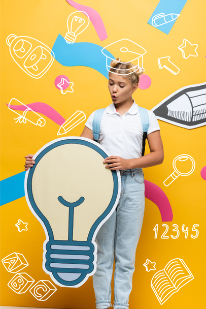 изумленный ученик держит светлый пузырь макет рядом с разноцветными элементами, бумажный карандаш и иллюстрации на желтом - Фото, изображение
