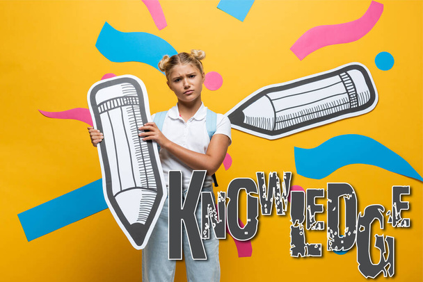 Sad schoolgirl holding paper pencil and looking at camera near knowledge lettering on yellow background - Fotoğraf, Görsel