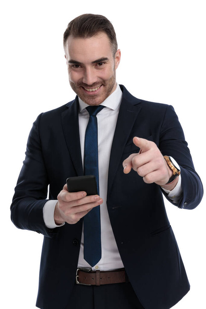 Happy businessman pointing and holding phone, smiling while standing on white studio background - Photo, Image
