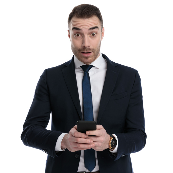 Удивленный бизнесмен улыбается и держит телефон, стоя на белом фоне студии - Фото, изображение