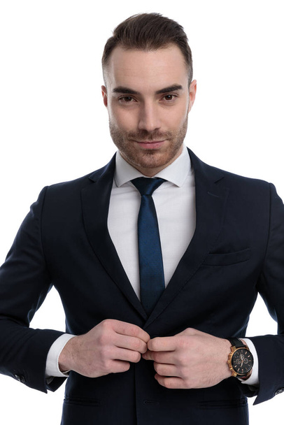 Selbstbewusster Geschäftsmann lächelt und justiert seine Jacke, während er auf weißem Studiohintergrund steht - Foto, Bild